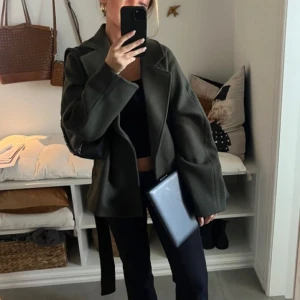 grön kappa från Gina Tricot - Stilren grön kappa från Gina Tricot med bred krage och bälte i midjan. Jackan har lång ärm och en klassisk, rak passform som ger en snygg siluett. Perfekt för dig som vill ha en enkel men trendig look under höst och vinter.