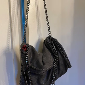 Grå axelväska med kedjedetaljer - Snygg grå shimrig axelväska med grova silverfärgade kedjor längs kanterna och som axelrem. Väskan är gjord i ett glansigt mockaliknande material och har röd insida med dragkedjefack. Stella McCartney liknande, fint skick (intresse koll)