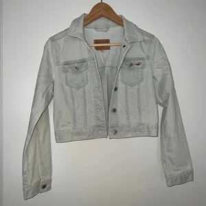 Ljus bomull jeansjacka från Hollister  - Snygg ljus jeansjacka från Hollister i klassisk modell med knappar framtill och två bröstfickor. Jackan är croppad och har långa ärmar. Perfekt att slänga över en hoodie eller t-shirt för en chill look. Har ett litet rosa märke som man ser på bilden men absolut inget som syns i sin helhet 