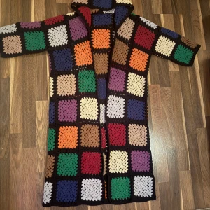 Färgglad virkade kappa  - Unik och färgglad lång cardigan jacka med virkade rutor i olika färger som grönt, lila, blått, rött, orange och beige. Jackan har lång ärm och öppen framtill, perfekt för dig som gillar retro och boho vibes. Supermysig att slänga på sig när det är lite kyligt.