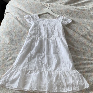 Vit sommarklänning från H&M - Superfin vit klänning från H&M i 100% bomull med broderade blommor och volang nertill. Den har korta ärmar och en luftig passform, perfekt för varma dagar. Söt och enkel design som känns fräsch och somrig.