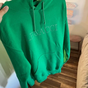 Grön hoodie från Legacci med strass - Snygg grön hoodie från Legacci med strassdetaljer på bröstet och en stor fjäril i strass på ryggen. Tröjan har huva med snörning och en klassisk känguruficka framtill. Perfekt för dig som vill sticka ut med coola detaljer.