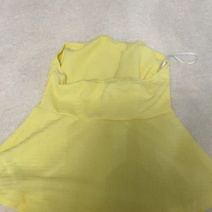 Gul bandeau tubtopp i lätt tyg - Snygg gul bandeau tubtopp med lätt, något transparent tyg och utsvängd nederdel. Toppen är axelbandslös och har en enkel, stilren design som passar perfekt till varma dagar. Perfekt för dig som vill ha en färgklick i garderoben!