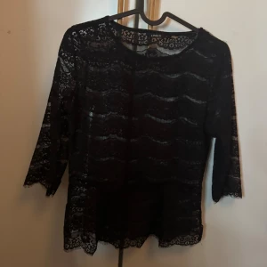 Svart spetsblus från Lindex XS - Svart blus från Lindex i storlek XS, helt i genombruten spets med vågigt mönster. Blusen har trekvartsärm och rund halsringning. Perfekt för dig som gillar en feminin och trendig look med snygga spetsdetaljer.