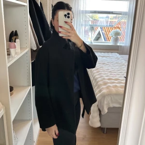 Svart kappa med bälte - Snygg svart kappa med bälte i midjan och långa ärmar. Klassisk rak modell som ger en clean och stilren look. Perfekt för dig som gillar minimalistisk stil och vill ha en jacka som funkar till många outfits.