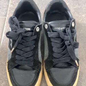 Zadig & Voltaire svarta sneakers - Snygga svarta sneakers från Zadig & Voltaire med cool blixtdetalj på sidan. Skorna har svart mocka och skinn, silverfärgade metalldetaljer och snörning. Sidan är i glittrigt tyg som ger extra edge. Priset kan diskuteras 