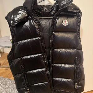  Moncler väst  - Snygg svart dunväst från Moncler med glansig finish och hög krage. Västen har två dragkedjefickor framtill och tydlig Moncler-logga på bröstet. Perfekt att slänga över en hoodie för en cool look.