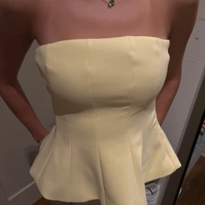 Gul topp från Zara - Säljer en ljusgul topp från Zara i storlek S. Toppen är axelbandslös med bandeauform och har en snygg utsvängd siluett nedtill. Materialet känns som polyester och toppen har markerade sömmar framtill för en strukturerad look. Aldrig använd 