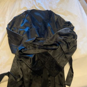Svart omlottjacka i satin - Snygg svart omlottjacka i glansigt satinmaterial med breda ärmar och knytband i midjan. Morgonrocken har en elegant känsla och spetsdetaljer längs kanten som ger en extra touch. Perfekt för dig som vill ha något stilrent och trendigt.