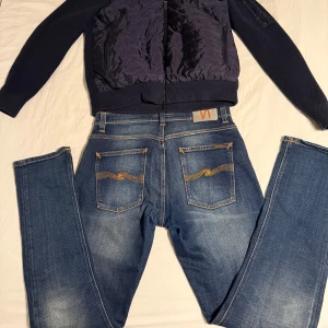 Nudie Jeans Grim Tim 33/32 - Säljer ett par klassiska blå jeans från Nudie Jeans Co i storlek 33/32. Jeansen har snygga slitningar, orange kontrastsömmar och ikoniska bakfickor med vågig brodyr. Tillverkade i bomull med knappgylf och normal passform. Perfekta för en avslappnad streetstil. Hör av er vid frågor!