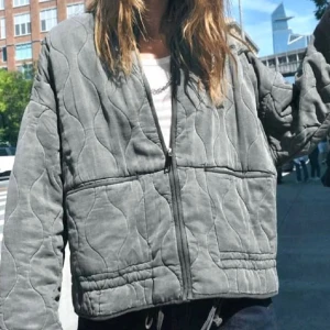 Grå quiltad jacka  - Säljer en grå quiltad bomberjacka med vågigt mönster och dragkedja framtill. Jackan har en loose passform och långa ärmar, perfekt för dig som gillar en avslappnad streetstil. Den har 2 fläckar där färgen har försvunnit!!
