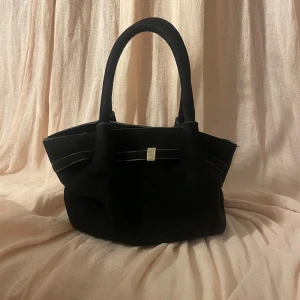 Svart mocka handväska med gulddetalj - Stilren svart handväska i mjuk mocka med två rejäla handtag och en diskret guldfärgad detalj framtill. Väskan har en klassisk form och passar perfekt för dig som gillar en clean och elegant look. 