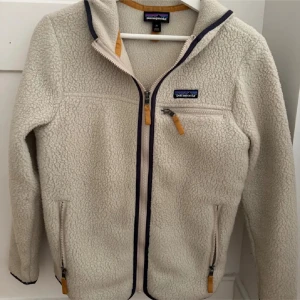 Beige teddyjacka från Patagonia XS - Mysig beige teddyjacka från Patagonia i storlek XS. Jackan har huva, hel dragkedja framtill, tre fickor med dragkedja och detaljer i mörkblått och gult. 