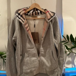 Burberry Tröja - Snygg grå tröja ifrån Burberry. Klassisk huva med Burberrys rutiga mönster på insidan och broderad logga på bröstet.