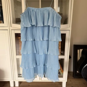 Ljusblå volangklänning med axelband - Superfin ljusblå klänning med tunna axelband och flera lager volanger. Plisserad struktur ger ett luftigt och somrigt intryck. Perfekt för dig som gillar romantisk och trendig stil.