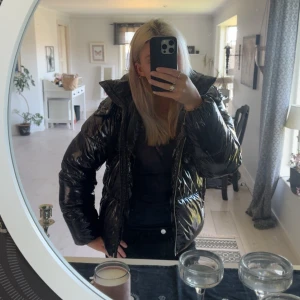 Svart glansig pufferjacka från Nelly - Säljer en svart, glansig pufferjacka från Nelly med huva och dragkedja. Använd 1 gång, nypris ca 800🩷