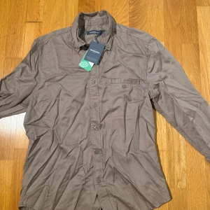 Brun skjorta från Dressmann - Säljer en brun långärmad skjorta från Dressmann i klassisk modell. Skjortan har knappar hela vägen, bröstficka och stilren krage. Perfekt för dig som gillar en enkel och clean look. Materialet känns mjukt och lätt, passar till många olika outfits. Behövs strykas