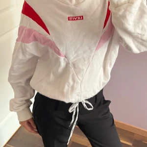 Vit och röd sweatshirt från Levi's XS - Säljer en snygg vit sweatshirt från Levi's i storlek XS med röda och rosa detaljer i ett coolt mönster över bröstet och ärmarna. Tröjan har lång ärm, snörning med Levi's-logga i midjan och är gjord i mjuk bomull. Perfekt för dig som gillar streetstyle och färg.