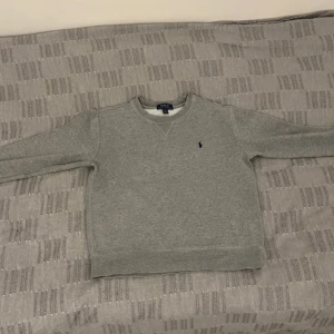 Grå sweatshirt från Polo Ralph Lauren - Klassisk grå sweatshirt från Polo Ralph Lauren med rund halsringning och broderad marinblå logga på bröstet. Tröjan har ribbade muddar vid ärmslut och nederkant, perfekt för en chill och stilren look.