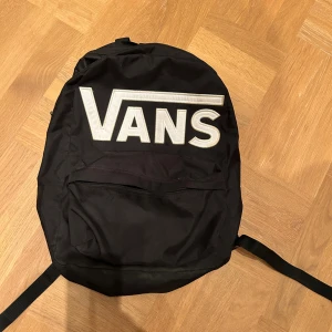 Svart ryggsäck från Vans - Säljer en svart ryggsäck från Vans med stort vitt logotryck på framsidan. Väskan har ett stort huvudfack och en mindre ficka framtill med dragkedja. Justerbara axelremmar och klassisk form, perfekt för skolan eller stan.