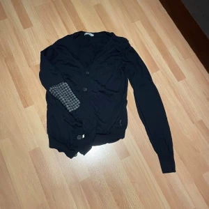 Navy blå kofta från J.Lindeberg med rutiga detaljer - Navy blå kofta från J.Lindeberg med knäppning framtill och rutiga grå armbågslappar. Långärmad modell i mjuk bomull, perfekt för lager-på-lager. Klassisk och stilren med en twist tack vare de rutiga detaljerna på ärmarna.