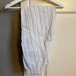 Randiga vita pyjamasbyxor med snörning - Snygga vita pyjamasbyxor med svarta tunna ränder och elastisk midja med snörning. Byxorna har avslappnad passform och raka ben, perfekta för en chill och stilren look. Materialet är lätt och luftigt, vilket gör dem grymma för varmare dagar.