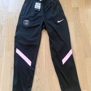 Svarta PSG träningsbyxor från Nike - Svarta träningsbyxor från Nike med PSG-logga på ena benet och rosa detaljer på båda benen. Byxorna har elastisk midja och är gjorda i Dri-Fit polyester som andas. Perfekta för fotboll och sportiga aktiviteter.