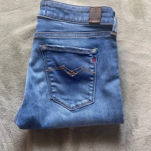 Replay blå slim jeans - Säljer ett par blå jeans från Replay med klassisk femficksdesign och snygga detaljer på bakfickan. Jeansen har en slim passform och är tillverkade i mjukt denimtyg med lätt tvättad look. Perfekta för dig som gillar stilrena och bekväma jeans.