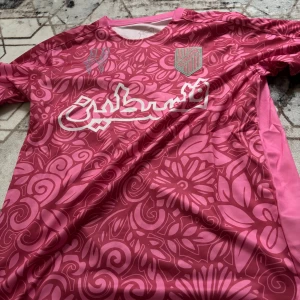 FC Palestine rosa blommig fotbollströja. Helt ny - Snygg fotbollströja från FC Palestine i rosa med blommigt mönster och vit text på bröstet. Kortärmad modell i lätt och ventilerande polyester, perfekt för träning eller match. Unik design med klubbmärke broderat på vänster sida.