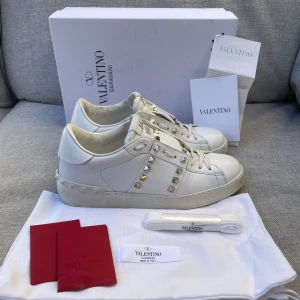 Valentino skor  - Säljer ett par vita Valentino Garavani Rockstud sneakers i skinn med ikoniska guld- och silverfärgade nitar. Klassisk rund tå, snörning och platt sula. Kommer med originalkartong, extra skosnören och dustbag. Perfekta för dig som vill sticka ut med lyxig streetstyle. Dustbag medföljer bara!
