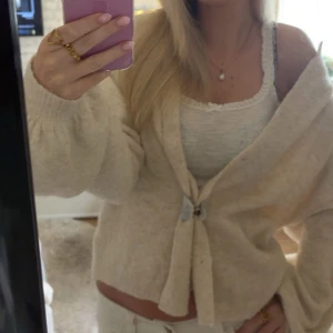 Beige stickad kofta - Supermysig beige stickad kofta med v-ringad hals och stora, fluffiga ärmar. Koftan har knappar framtill och en loose passform som ger en chill vibe. Perfekt att slänga över ett linne eller t-shirt för en snygg lager-på-lager look.