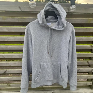 Grå hoodie från Sunspel med huva - Grå hoodie från Sunspel i storlek S, tillverkad i mjuk bomull. Tröjan har en stor huva med snörning, klassisk känguruficka framtill och ribbade muddar vid ärmslut och nederkant. Perfekt för chill dagar och enkel att matcha med jeans eller joggers.