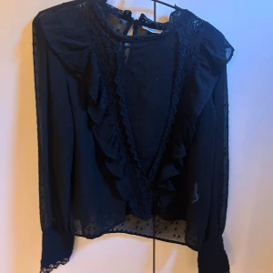 Svart blus med volang och spets - Säljer en svart blus med djup v-ringning framtill, dekorerad med volanger och spetsdetaljer. Blusen är transparent med diskret prickigt mönster i tyget och har långa ärmar med spets vid ärmslut. Knytning i nacken och lätt flowy passform.