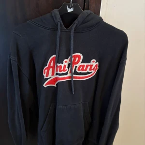 Svart hoodie Ami Paris - Snygg svart hoodie från Ami Paris med stort rött och vitt broderat tryck på bröstet. Klassisk huva med snörning och magficka framtill. Perfekt för dig som gillar fashion! Skriv vid frågor osv