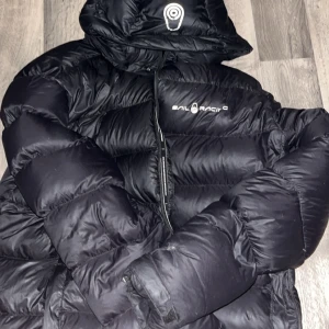 Svart pufferjacka från Sail Racing XL - Säljer en svart pufferjacka från Sail Racing i storlek XL. Jackan har huva med logga, tydlig Sail Racing-tryck på bröstet och grova, quiltade sektioner. Materialet är syntet och jackan har dragkedja framtill. Perfekt för kalla dagar och riktigt snygg