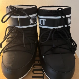 Svarta Moon Boot snow boots - Svarta Moon Boot snow boots med vit logga och snörning framtill. Skorna har en rund tå, platt sula och är tillverkade i syntetmaterial. Perfekta för vintern med sin ikoniska design och höga komfort. Snygga och praktiska för kalla dagar. Har använt dom kanske 3-5 gånger förra vintern. Jag bor där de inte är så kalt så skorna kom aldrig till användning. Har påsen till skorna kvar och lådan.