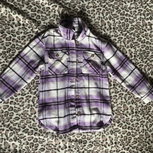 Lila rutig overshirt  - Snygg jacka/skjorta från Lindex i lila, vit och svart rutig design. Skjortan har två bröstfickor med knappar, klassisk krage och långa ärmar. Perfekt att slänga över en t-shirt för en chill look. Skönt och tjockt tyg
