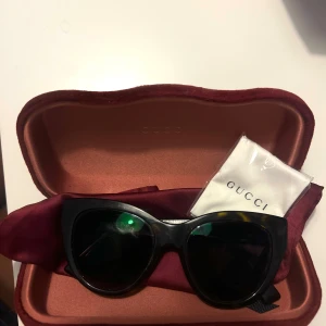 Svarta cat-eye solglasögon från Gucci - Snygga svarta cat-eye solglasögon från Gucci med tjock båge och diskret logga på sidorna.                                  Haft i 1 år använder knappt dem dem har några få repor på sig så man är medveten.  Köpta på synsam Originellt pris är 2229 . Solglasögonen är brun och svart. Kommer med original fodralet.