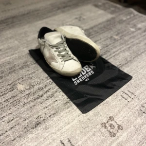 Golden Goose Superstar  - Tjenare! Säljer nu ett par Golden Goose Superstar sneakers i vitt skinn med grå mockadetaljer och svart häl. Klassisk stjärndesign på sidan, grå snörning och platt sula. Snygg och trendig modell som passar perfekt till streetwear och casual outfits. Skorna har bara andvänds några få tal gånger. Det är bara att fråga vid ytterligare frågor! 