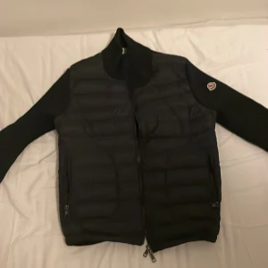 Svart cardigan från Moncler - Svart cardigan från Moncler med quiltad framsida och tvåvägsdragkedja. Cardigan har hög krage och Moncler-logga på vänster axel. Materialmix med dun och släta partier ger en clean och sportig look.