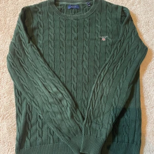 Grön kabelstickad tröja från Gant - Mörkgrön kabelstickad tröja från Gant i 100% bomull. Klassisk rund halsringning, ribbade muddar och broderad Gant-logga på bröstet. Perfekt för lager-på-lager och snygg till jeans eller chinos.
