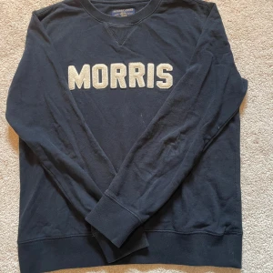 Mörkblå sweatshirt från Morris - Mörkblå sweatshirt från Morris med stora, broderade bokstäver i vitt framtill. Klassisk rund halsringning, ribbade muddar och lång ärm. Tillverkad i mjuk bomull, perfekt för en chill och stilren look.