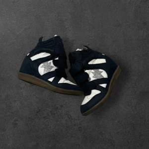 Svartvita sneakers med kilklack - Snygga svartvita sneakers med kilklack och kardborreband. Skorna har en cool silverglittrig stjärndetalj på sidan och en brun sula. Ovandelen är i mocka och syntet, vilket ger en sportig men ändå trendig look. Perfekta för dig som vill ha lite extra höjd och stil.