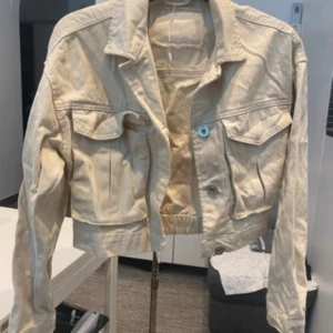 Beige croppad jeansjacka - Snygg beige croppad jeansjacka med klassisk krage, två bröstfickor med lock och knappar framtill. Jackan har långa ärmar och elastisk midja för en modern passform. Perfekt för dig som vill ha en stilren och trendig look.