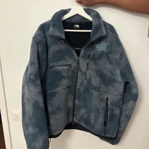 Blå fleecejacka från The North Face - Mysig blå fleecejacka från The North Face med broderad logga på bröstet och ryggen. Jackan har flera dragkedjeförsedda fickor och hög krage. Perfekt för kyliga dagar och har en riktigt skön, fluffig känsla.