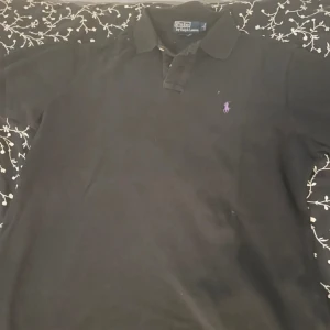 Svart pikétröja från Ralph Lauren - Klassisk svart pikétröja från Polo Ralph Lauren med lila broderad logga på bröstet. Tröjan har krage, korta ärmar och knäppning framtill. Perfekt för en stilren och avslappnad look.