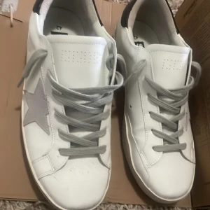 Golden Goose Superstar sneakers vita/grå - Golden Goose Superstar sneakers i vitt skinn med grå mockastjärna på sidan, grå snörning och svart detalj på hälen. Klassisk rund tå och platt sula med vintagekänsla. Perfekta för dig som gillar streetstyle och snygga detaljer.