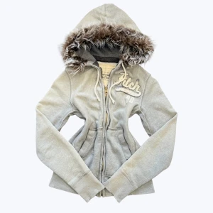 Grå hoodiejacka med fuskpäls från Abercrombie - Grå hoodiejacka från Abercrombie & Fitch med dragkedja, broderad logga på bröstet och mysig huva med fuskpäls. Jackan har långa ärmar, ribbade muddar och två fickor framtill. Perfekt för kyliga dagar när du vill vara både bekväm och snygg. Inga prisförlsag‼️