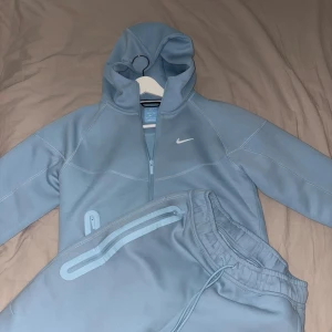 Nike NOCTA tech fleece - Snygg ljusblå hoodie från Nike NOCTA med huva och dragkedja framtill. Tröjan har vita logodetaljer på bröstet och ryggen, samt stilrena sömmar som ger en sportig vibe. Perfekt för chill eller träning, och matchar lätt med andra plagg.