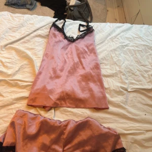 Dusty rose Rosa satin pyjamas med svart spetsdetalj - Säljer ett rosa pyjamas sett i satin med svarta spetsdetaljer längs v-ringningen och tunna axelband.  Toppen har även en  glansig finish och en feminin look, perfekt för att lyfta vilken outfit som helst. Supersnygg att styla med jeans eller kjol. Denna Pyjamasen är stilren och ger den rätta viben.